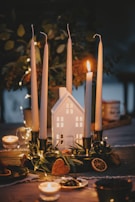 A cozy arrangement featuring a centerpiece with four tall candles placed in candle holders around a small house-shaped ornament. The candles are unlit except for one, creating a warm glow. Surrounding the base of the candles are various decorative elements like dried orange slices, green foliage, and a heart-shaped item. The background is softly blurred, with twinkling fairy lights adding to the ambient atmosphere.