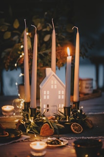 A cozy arrangement featuring a centerpiece with four tall candles placed in candle holders around a small house-shaped ornament. The candles are unlit except for one, creating a warm glow. Surrounding the base of the candles are various decorative elements like dried orange slices, green foliage, and a heart-shaped item. The background is softly blurred, with twinkling fairy lights adding to the ambient atmosphere.