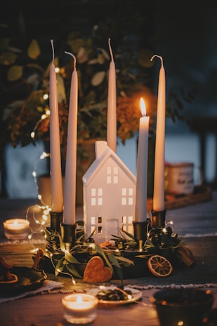 A cozy arrangement featuring a centerpiece with four tall candles placed in candle holders around a small house-shaped ornament. The candles are unlit except for one, creating a warm glow. Surrounding the base of the candles are various decorative elements like dried orange slices, green foliage, and a heart-shaped item. The background is softly blurred, with twinkling fairy lights adding to the ambient atmosphere.