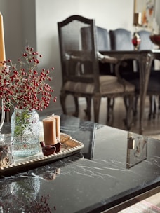 A sophisticated dining area features a glossy black marble table with a decorative tray holding two tall candles and a vase filled with small red berries. In the background, a classic wooden dining set with cushioned chairs adds elegance.