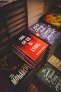 Stacks of books with colorful and intriguing covers are neatly arranged. Titles such as 'One of Us is Lying', 'The Monarchs', and 'We'll Always Have Summer' are prominently displayed. The scene suggests a bookstore or a book fair setting.