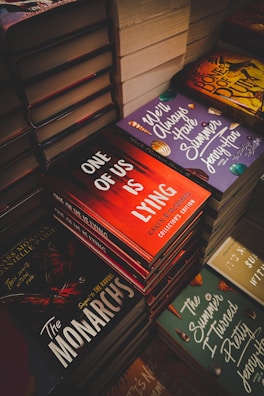 Stacks of books with colorful and intriguing covers are neatly arranged. Titles such as 'One of Us is Lying', 'The Monarchs', and 'We'll Always Have Summer' are prominently displayed. The scene suggests a bookstore or a book fair setting.