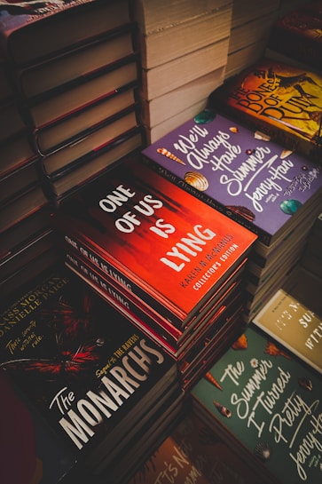 Stacks of books with colorful and intriguing covers are neatly arranged. Titles such as 'One of Us is Lying', 'The Monarchs', and 'We'll Always Have Summer' are prominently displayed. The scene suggests a bookstore or a book fair setting.