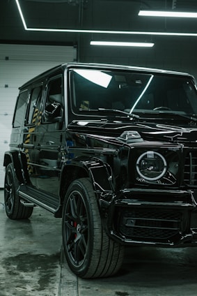 A luxury black SUV is parked indoors, surrounded by a modern industrial setting with sleek black and metallic surfaces. Overhead lights create reflective highlights on the vehicle's glossy surface, and the large tires and distinctive front grille are prominently displayed.