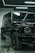 A luxury black SUV is parked indoors, surrounded by a modern industrial setting with sleek black and metallic surfaces. Overhead lights create reflective highlights on the vehicle's glossy surface, and the large tires and distinctive front grille are prominently displayed.