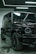 A luxury black SUV is parked indoors, surrounded by a modern industrial setting with sleek black and metallic surfaces. Overhead lights create reflective highlights on the vehicle's glossy surface, and the large tires and distinctive front grille are prominently displayed.