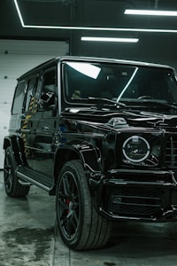 A luxury black SUV is parked indoors, surrounded by a modern industrial setting with sleek black and metallic surfaces. Overhead lights create reflective highlights on the vehicle's glossy surface, and the large tires and distinctive front grille are prominently displayed.