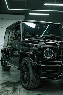 A luxury black SUV is parked indoors, surrounded by a modern industrial setting with sleek black and metallic surfaces. Overhead lights create reflective highlights on the vehicle's glossy surface, and the large tires and distinctive front grille are prominently displayed.
