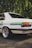 A vintage white car is parked on a cobblestone surface, with a green and blue stripe running along its side. The car features shiny chrome rims and a classic design with reflector tail lights. Lush green foliage provides a natural backdrop, creating a serene outdoor setting.