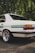 A vintage white car is parked on a cobblestone surface, with a green and blue stripe running along its side. The car features shiny chrome rims and a classic design with reflector tail lights. Lush green foliage provides a natural backdrop, creating a serene outdoor setting.