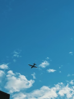 A large aircraft is flying high in the sky, surrounded by scattered, fluffy white clouds. The sky is a vibrant blue, creating a stark contrast with the dark silhouette of the plane.