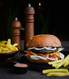 A juicy hamburger is topped with lettuce, onion rings, pickles, and tomatoes, all sandwiched in a sesame seed bun. It is accompanied by a side of crispy golden fries, two small black dishes containing different sauces, and two tall wooden pepper mills in the background. The setup is on a dark surface, creating a contrast with the vibrant colors of the food.