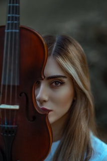 A person with long hair is partially obscured by a violin. The violin is held upright, covering half of the face, and has a rich, dark brown finish. The person's eyes are visible and focus directly at the camera. The background is blurred with earthy tones.