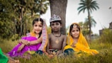 Three children are dressed in traditional attire, sitting on the grass against a backdrop of trees. The child in the center wears a crown and is shirtless, while the two children on either side are in colorful clothing and adorned with jewelry.