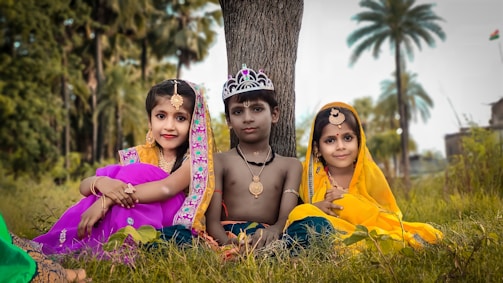 Three children are dressed in traditional attire, sitting on the grass against a backdrop of trees. The child in the center wears a crown and is shirtless, while the two children on either side are in colorful clothing and adorned with jewelry.