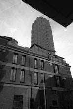 A tall, modern skyscraper rises above older brick buildings, with the word 'Sumatra' visible on one. The scene is depicted in black and white, and a few people can be seen walking at street level. The sky appears slightly overcast.