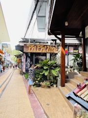 A small street with various shops and stalls on either side. A person stands near a wooden sign that reads 'Phi Phi Inn,' surrounded by potted plants and tropical greenery. The walkway is paved with intricate tile designs, and there are people in the background exploring the area.