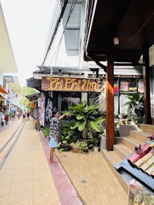 A small street with various shops and stalls on either side. A person stands near a wooden sign that reads 'Phi Phi Inn,' surrounded by potted plants and tropical greenery. The walkway is paved with intricate tile designs, and there are people in the background exploring the area.