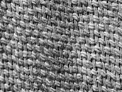 A close-up view of a woven fabric texture showcasing the intricate interlacing of thick, fibrous threads. The black and white image highlights the rough, tactile surface and the complex pattern of the weave.