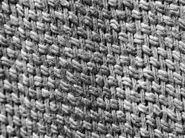 A close-up view of a woven fabric texture showcasing the intricate interlacing of thick, fibrous threads. The black and white image highlights the rough, tactile surface and the complex pattern of the weave.
