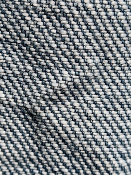 Close-up view of a woven textile pattern, featuring tightly interlaced threads that create a textured surface. The material displays a diagonal weave with contrasting colors of dark blue and light beige.