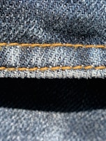 Close-up view of denim fabric highlighting the texture of the woven material. The stitching is a prominent feature, showing a line of neat orange thread running across the fabric.