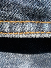 Close-up view of denim fabric highlighting the texture of the woven material. The stitching is a prominent feature, showing a line of neat orange thread running across the fabric.