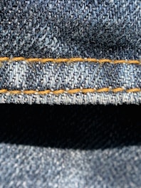 Close-up view of denim fabric highlighting the texture of the woven material. The stitching is a prominent feature, showing a line of neat orange thread running across the fabric.