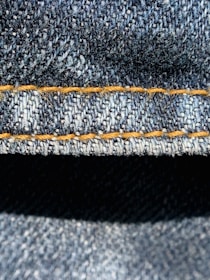Close-up view of denim fabric highlighting the texture of the woven material. The stitching is a prominent feature, showing a line of neat orange thread running across the fabric.