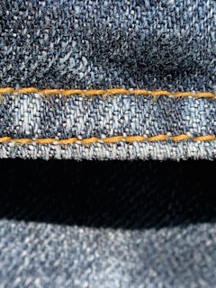 Close-up view of denim fabric highlighting the texture of the woven material. The stitching is a prominent feature, showing a line of neat orange thread running across the fabric.