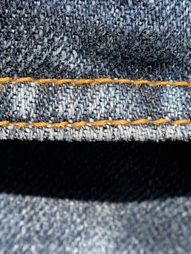 Close-up view of denim fabric highlighting the texture of the woven material. The stitching is a prominent feature, showing a line of neat orange thread running across the fabric.