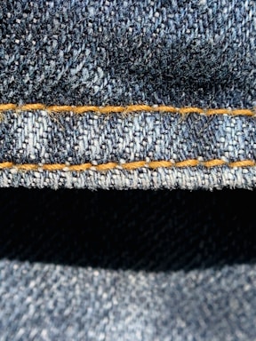 Close-up view of denim fabric highlighting the texture of the woven material. The stitching is a prominent feature, showing a line of neat orange thread running across the fabric.