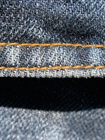 Close-up view of denim fabric highlighting the texture of the woven material. The stitching is a prominent feature, showing a line of neat orange thread running across the fabric.