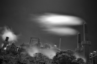 An industrial facility with large chimneys releasing smoke into the sky, surrounded by tall structures and machinery. The night setting creates a stark contrast between the lit smoke plumes and the dark sky. Vegetation in the foreground appears somewhat blurred, possibly due to motion or exposure settings.