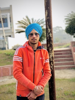 A person stands outdoors in front of a pole, wearing an orange Adidas track jacket and a blue turban. Sunglasses cover their eyes, and a building and greenery are visible in the background.