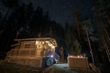 A cozy wooden cabin is nestled in a forest at night, warmly lit by string lights that adorn the porch. A hot tub is positioned nearby, emitting steam, set against a backdrop of tall trees and a starlit sky.