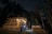 A cozy wooden cabin is nestled in a forest at night, warmly lit by string lights that adorn the porch. A hot tub is positioned nearby, emitting steam, set against a backdrop of tall trees and a starlit sky.
