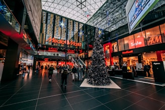 A bustling indoor shopping mall with festive Christmas decorations, including a large Christmas tree adorned with ornaments and stars. The mall features various store brands such as Media Markt and H&M, with sales signs visible. The high glass ceiling allows natural light to filter through, adding to the lively atmosphere.