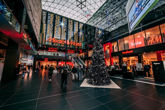 A bustling indoor shopping mall with festive Christmas decorations, including a large Christmas tree adorned with ornaments and stars. The mall features various store brands such as Media Markt and H&M, with sales signs visible. The high glass ceiling allows natural light to filter through, adding to the lively atmosphere.