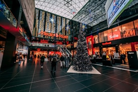 A bustling indoor shopping mall with festive Christmas decorations, including a large Christmas tree adorned with ornaments and stars. The mall features various store brands such as Media Markt and H&M, with sales signs visible. The high glass ceiling allows natural light to filter through, adding to the lively atmosphere.