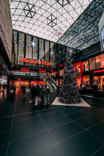 A large shopping mall interior features a festive Christmas tree decorated with ornaments. The ceiling is glass-panelled with star decorations, and the storefronts display bright signage, including 'Media Markt.' Shoppers walk by carrying bags, and the atmosphere is bustling with activity.