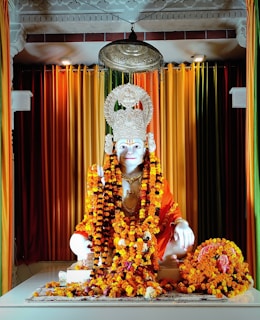 A white marble statue adorned with intricately designed garlands made of marigold flowers. The statue wears an ornate crown and is set against a backdrop of colorful drapes in hues of orange, yellow, green, and red. An ornate hanging lamp above adds a spiritual ambiance.
