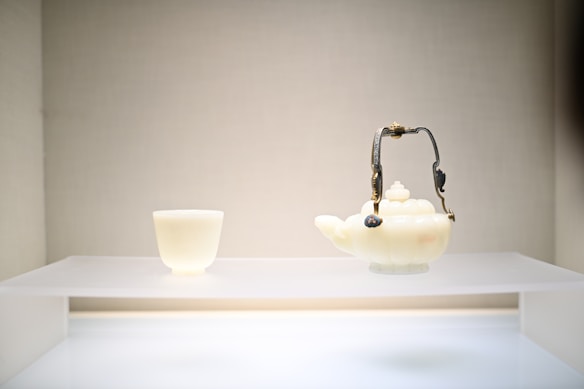 A white jade teapot with an ornate handle and an accompanying small cup are displayed on a white platform against a simple background.