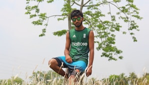A young man wearing sunglasses and athletic clothing is kneeling in a field. A tree with green leaves is in the background, and tall grass surrounds him.
