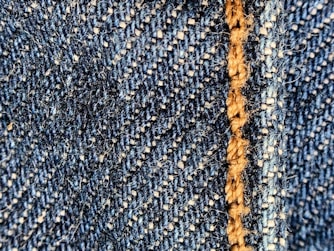 Close-up of a blue denim fabric with prominent stitching in light brown thread. The texture shows the interwoven fibers and a slightly fuzzy surface.