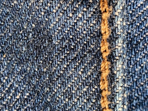 Close-up of a blue denim fabric with prominent stitching in light brown thread. The texture shows the interwoven fibers and a slightly fuzzy surface.