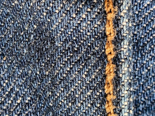 Close-up of a blue denim fabric with prominent stitching in light brown thread. The texture shows the interwoven fibers and a slightly fuzzy surface.