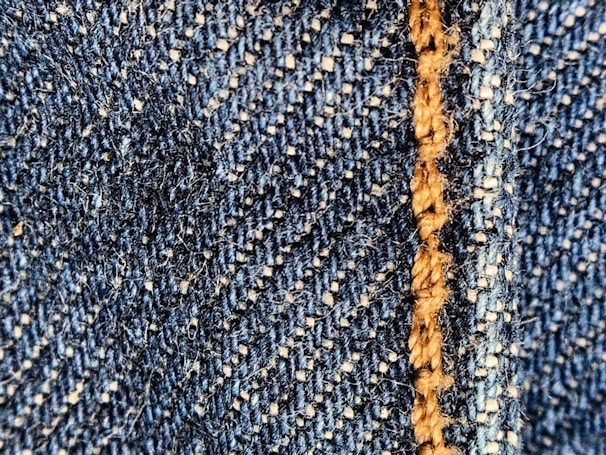 Close-up of a blue denim fabric with prominent stitching in light brown thread. The texture shows the interwoven fibers and a slightly fuzzy surface.