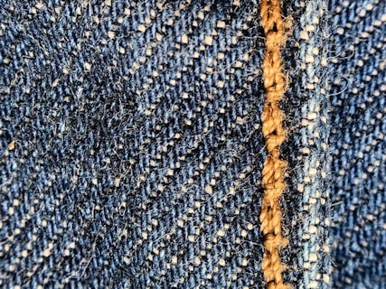 Close-up of a blue denim fabric with prominent stitching in light brown thread. The texture shows the interwoven fibers and a slightly fuzzy surface.