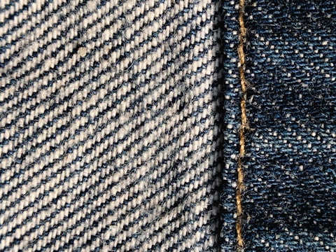 Close-up view of denim fabric showcasing a detailed weave pattern in blue and white. A seam with visible stitches runs vertically on the right side, highlighting the texture and craftsmanship.